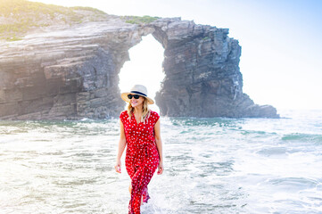 Blond woman wearing red dress and hat and walking along beach, Natural Arch at Playa de Las...