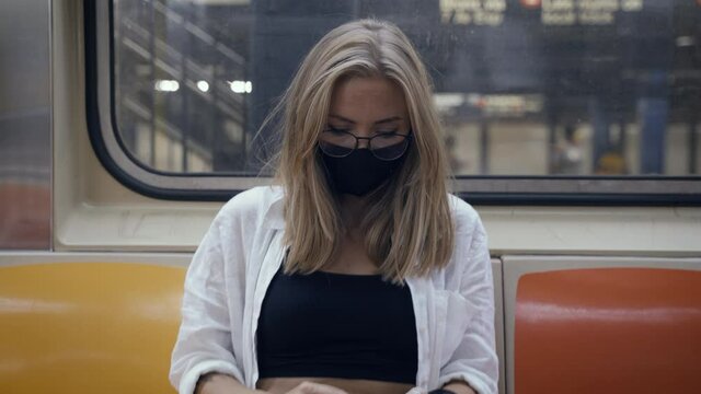 Portrait of a woman in a black medical face mask to avoid the spread of coronavirus who is sitting alone in a modern subway car. A girl in a surgical mask is keeping social distance on a metro train.