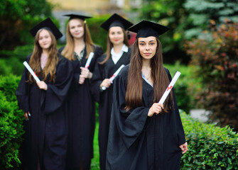 pretty young girl is a college graduate student in a black robe smiling holding a diploma in her hands against the background of a group of graduates.