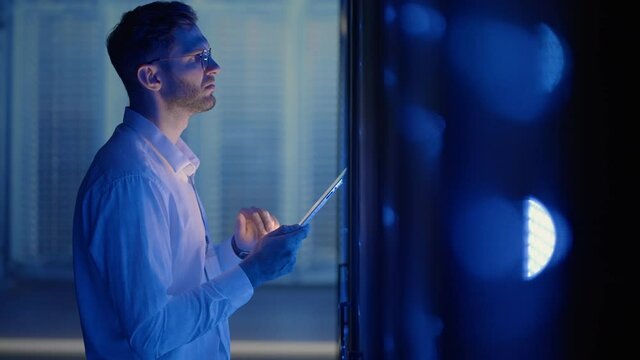 IT technician in data center digital server room controlling work of rack server cabinets with a tablet. Professional server engineer at work.