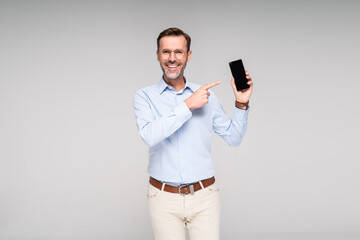 Smiling handsome man pointing on empty screen of smartphone, isolated on gray background.