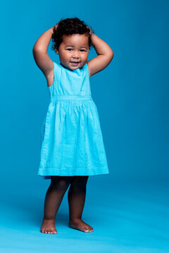Portrait of smiling little girl with hands on head wearing light blue dress standing against blue background