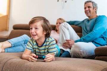 Boy lying on couch at home playing video game with parents watching
