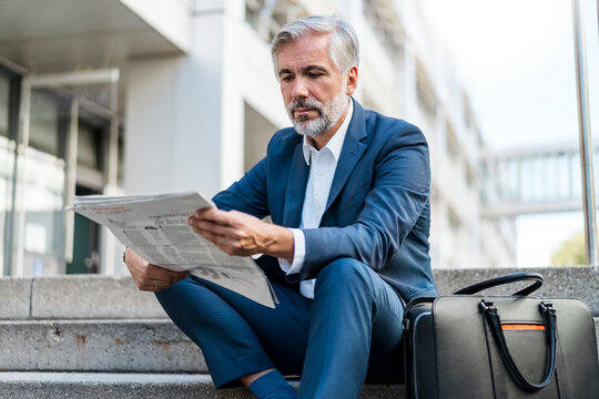 Mature businessman sitting on stairs in the city reading newspaper