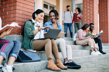 Happy multi-ethnic female students have fun while using laptop in front of university building.