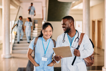 Happy multiracial medical students talk while reading medical record in hallway.