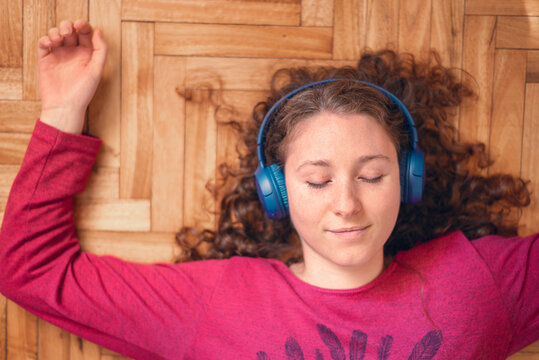 Top view of  young caucasian woman laying on the wooden floor with blue headphones. Relaxing at home. Smiling while listening to music with eyes closed