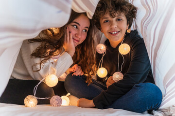 Portrait of happy brother and sister with chain of lights underneath bedcover