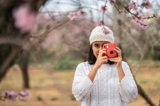 Portrait of teenage girl taking photo with camera among blossoming almond trees