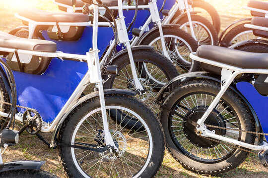 Many retro fat electric bicycle scooters parked on dirt grass field outdoor ready for hire rental sharing cpuntry parl leisure recreation trip. E power transport rent equipment. Rural offroad cycling