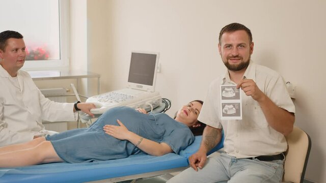 Caucasian male gynecologist giving images of future baby to happy husband after ultrasound scan of pregnant wife. Young family visiting doctor for medical examination during pregnancy.