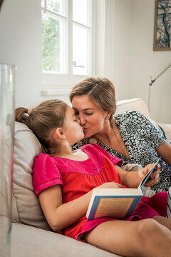 Mother kissing daughter with book on couch in living room