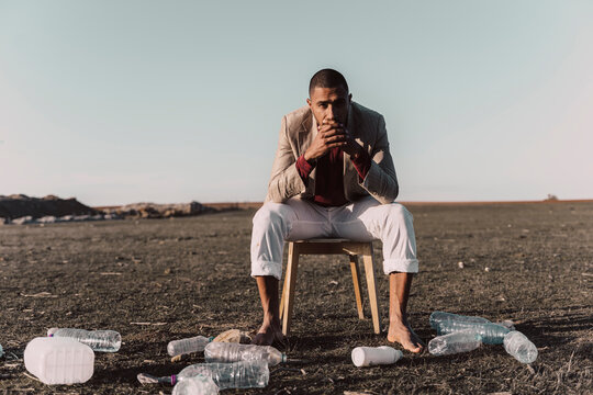 Young man sitting on chair surrounded by plastic bottles in barren land