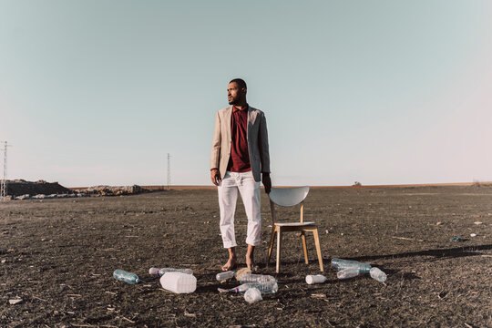 Young man standing next to chair surrounded by plastic bottles in barren land