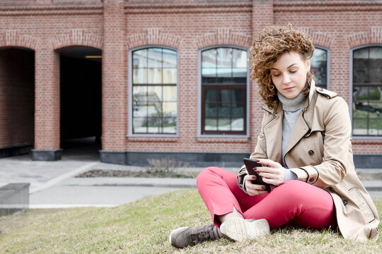Portrait of woman with curly hair sitting on a meadow looking at cell phone