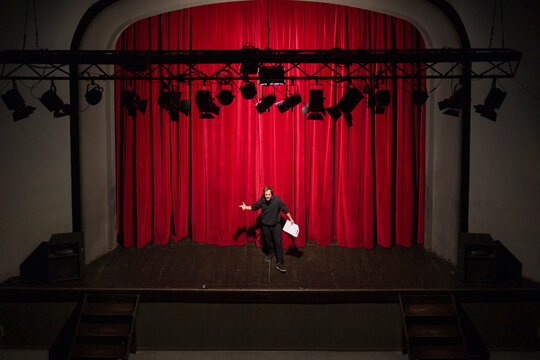 Rehearsing actor with script standing on theatre stage in front of red curtain