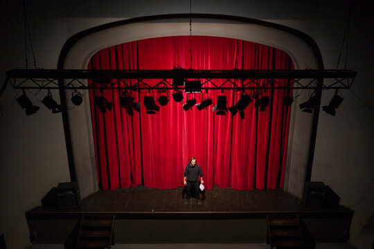 Actor with script standing on theatre stage in front of red curtain