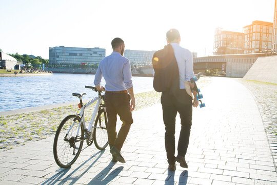 Two businessmen walking with bicycle and skateboard at the riverbank