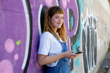 Smiling young woman using smartphone in front of purple graffiti wall