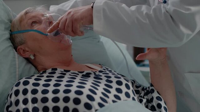 Doctor putting oxygen mask on face of hyperventilating patient with sickness. Close up of medic giving help to retired woman for healthcare treatment and recovery in hospital ward.
