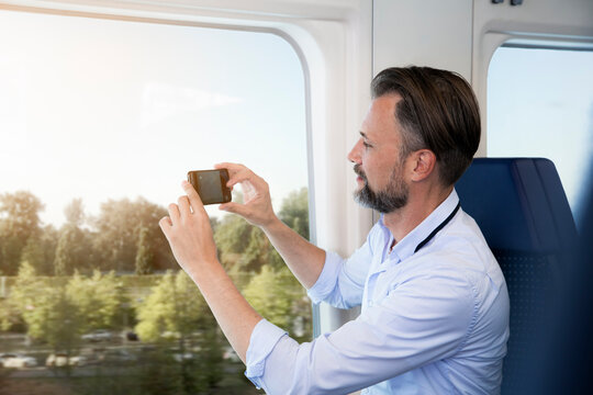 Mature man sitting in a train, taking pictures with his smartphone