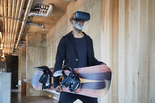 Mature businessman wearing VR glasses holding snowboard in modern office