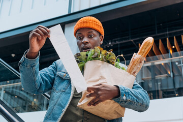 Doubting African-American person in denim jacket looks at sales paper receipt total holding pack with food products on escalator - obrazy, fototapety, plakaty