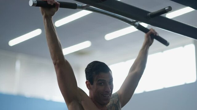 Positive confident fit Middle Eastern sportsman doing pull-ups smiling looking at camera. Happy self-assured handsome man working out training in gym posing. Endurance and bodybuilding