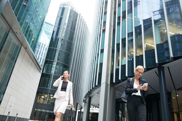 Female professionals walking with mobile phones against office building at downtown district in city