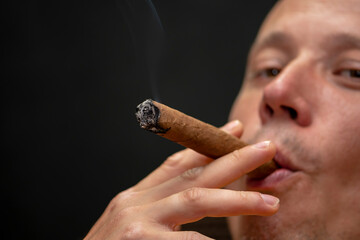 Portrait of a gray-haired man 40-45 years old on a dark background, smoking a cigar, sitting in an armchair, selective focus. Concept: a heavy smoker, the harm of tobacco, the cause of cancer.