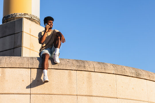 Young man talking over mobile phone while sitting on retaining wall against clear blue sky