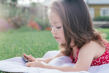 Portrait of little girl 3-4 in red lying on blanket on green grass and looking into mobile phone. Children using gadgets