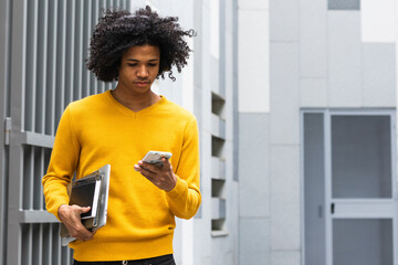 Curly hair boy with books using mobile phone while standing at university