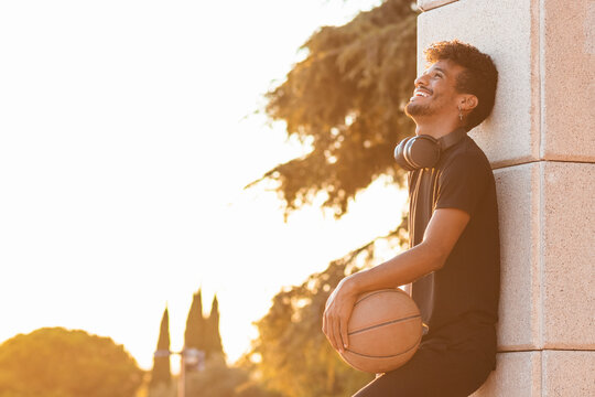 Cheerful young man holding basketball while standing by wall against clear sky during sunset