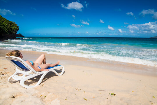 Woman relaxing on lounge chair at beach against blue sky, Grande Anse, Grenada, Caribbean