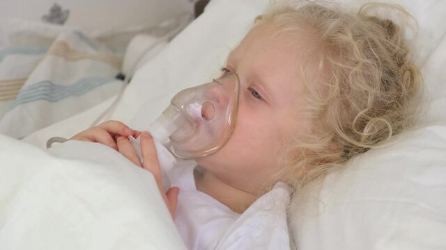cute little girl with curly blond hair lies in bed, sick five-tape girl in bed makes inhalation with a nebulizer and cryimg, treatment of bronchitis and lung disease in children