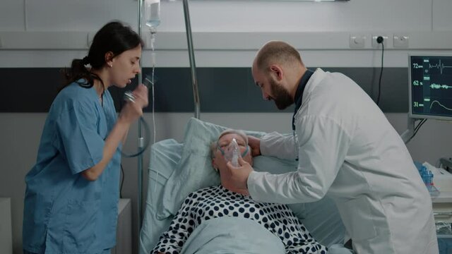 Doctor using oxygen tube mask for ill patient hyperventilating in hospital ward. Aged woman with heavy breathing problem receiving assistance from medic and nurse for healthcare.