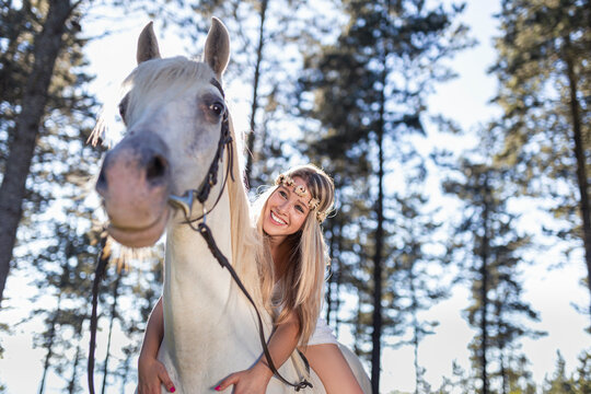 Low angle view of smiling young woman leaning on white horse in forest