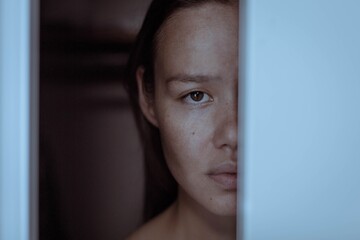 A sad face of a woman hiding behind a door afraid and terrified.