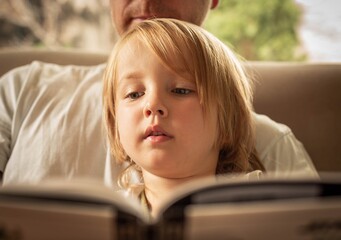 A little boy reading a book with his dad at home.