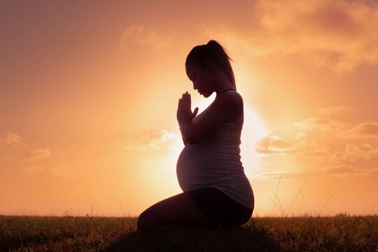 A pregnant woman sitting infront of a sunset with her hands folded in prayer.