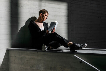Trans young man reading book while sitting on retaining wall in basement