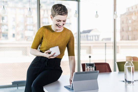 Smiling businesswoman sitting on desk in office holding coffee mug and using tablet - Powered by Adobe