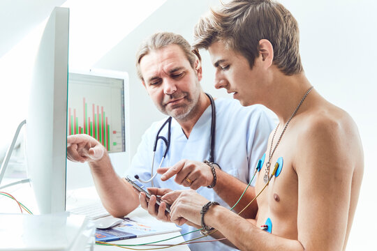 Doctor explaining to male shirtless patient with electrodes on chest over mobile phone in hospital