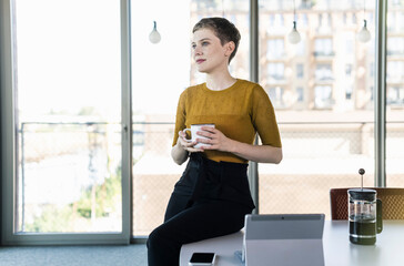 Businesswoman sitting on desk in office holding coffee mug