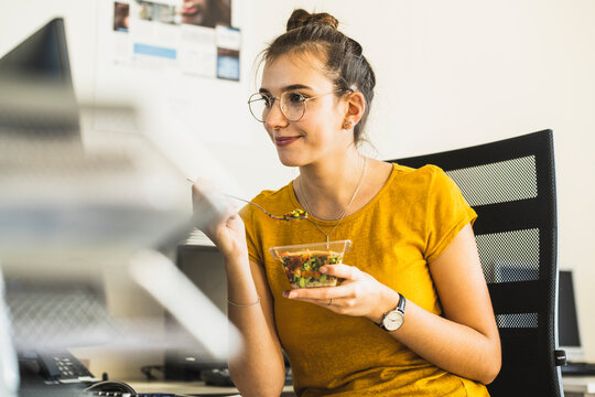 Smiling businesswoman eating food while looking at computer sitting at office
