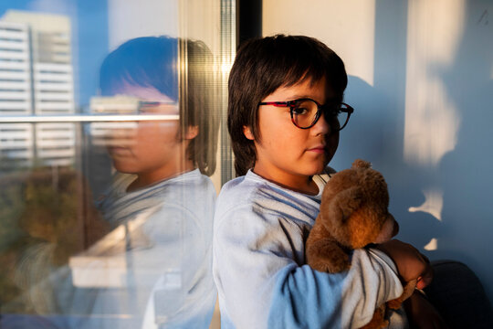 Portrait of serious boy with teddy bear leaning at window in the evening