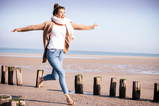 Happy young woman with arms outstretched running amidst wooden posts at beach during sunny day