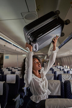 Young woman positioning luggage inside storage compartment in airplane