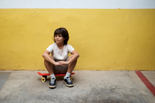 Thoughtful boy sitting on skateboard against wall
